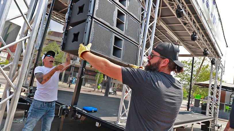 Derek Snowden, left, and Dana Akers, owners of Woolly Stage Company, raise a bank of speakers up Friday, May 19, 2023 as they set up their stage in front of the Springfield COhatch for IndieCraft 2023, which starts Friday evening and continues Saturday. BILL LACKEY/STAFF