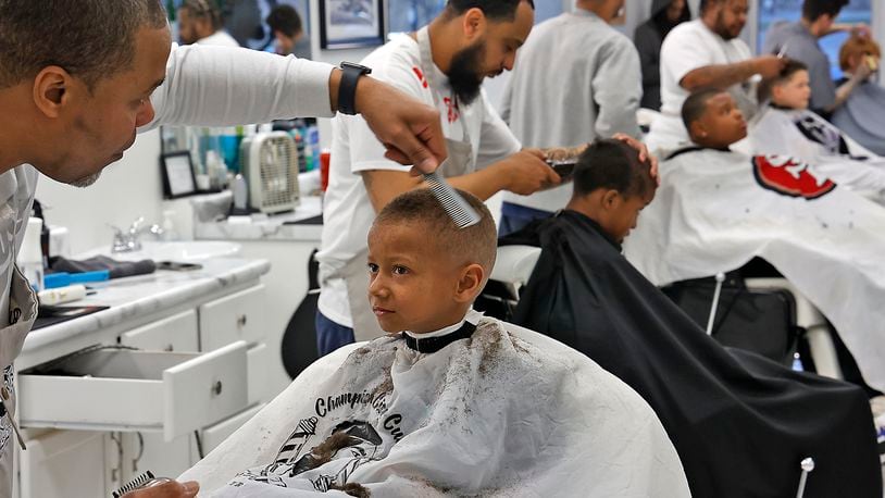 Maxx Johnson, 6, looks at Rufus Humphrey, one of the barbers at Champion City Cuts in the Southgate Shopping Center,  as he cuts his hair Monday, March 6, 2023.. The five barbers volunteer their time the first Monday of every month to give Springfield City School students of all ages a fresh cut and some pizza as they wait their turn in the chair. The haircuts are offered as an incentive by the school district each month for good attendance. BILL LACKEY/STAFF