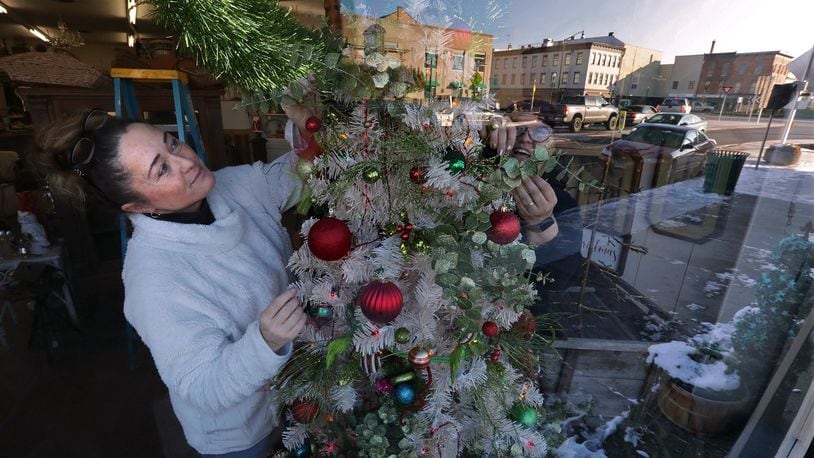 Downtown Urbana is reflected in the front window of the Vintage Traveler shop as owner, Tina Blakeman, and her friend Shannon Anderson decorate a Christmas tree in the window for the holidays earlier this month. BILL LACKEY/STAFF