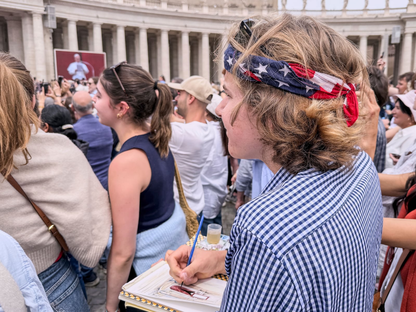 Notre Dame's architecture college student Wesley Buonerba, wearing a us flag bandanna, does a watercolor sketch of the loggia of St. Peter's Basilica during Pope Leo XIV first Sunday blessing of faithful gathered in the square for the traditional blessing, Sunday, May 11, 2025. (AP Photo/Giovanna Dell'Orto)