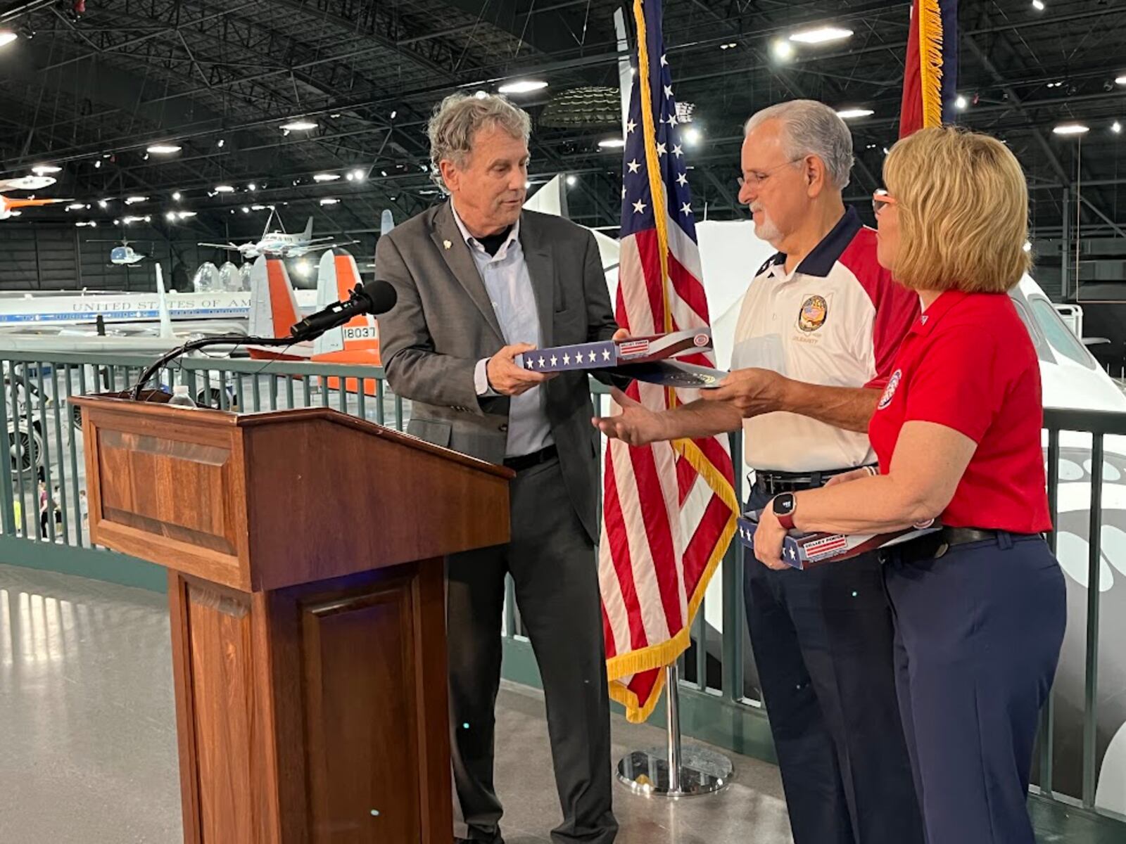U.S. Sen. Sherrod Brown, D-Ohio, hands Tom Ritchie, president of the Dayton-Miami Valley Regional Labor Council, a flag that has flown over the U.S. Capitol on Wednesday, Aug. 7, 2024, at the National Museum of the U.S. Air Force. Next to Ritchie is Kim Frisco, executive director of the Montgomery County Veterans Services Commission. THOMAS GNAU/STAFF