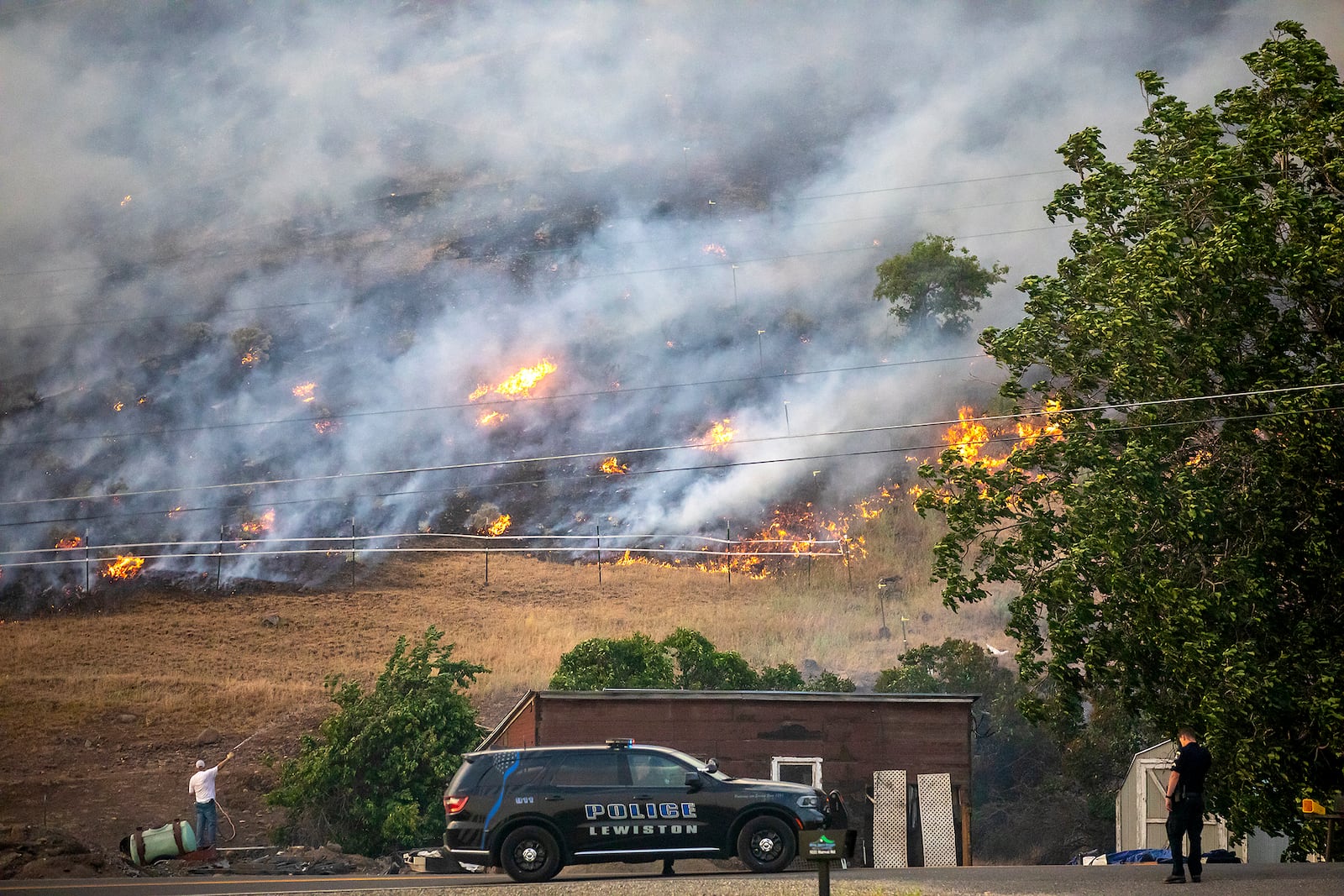 Lewiston police look on as a person uses a hose to keep the ground damp as strong winds push flames along the hills nearby, Saturday, May 31, 2025, in north Lewiston, Idaho. (August Frank/Lewiston Tribune via AP)