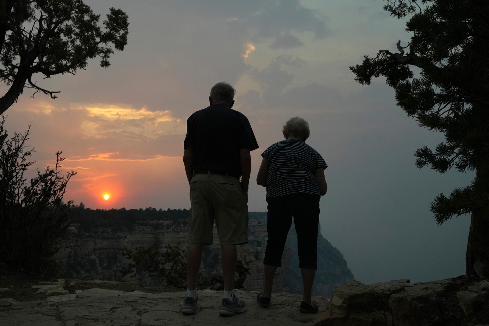 The setting sun peeks through the smoke from the Dragon Bravo Fire as it moves over the Grand Canyon from the North Rim as two tourists take in the scene from a South Rim overlook Monday, July 14, 2025, in Grand Canyon National Park, Ariz. (AP Photo/Ross D. Franklin)