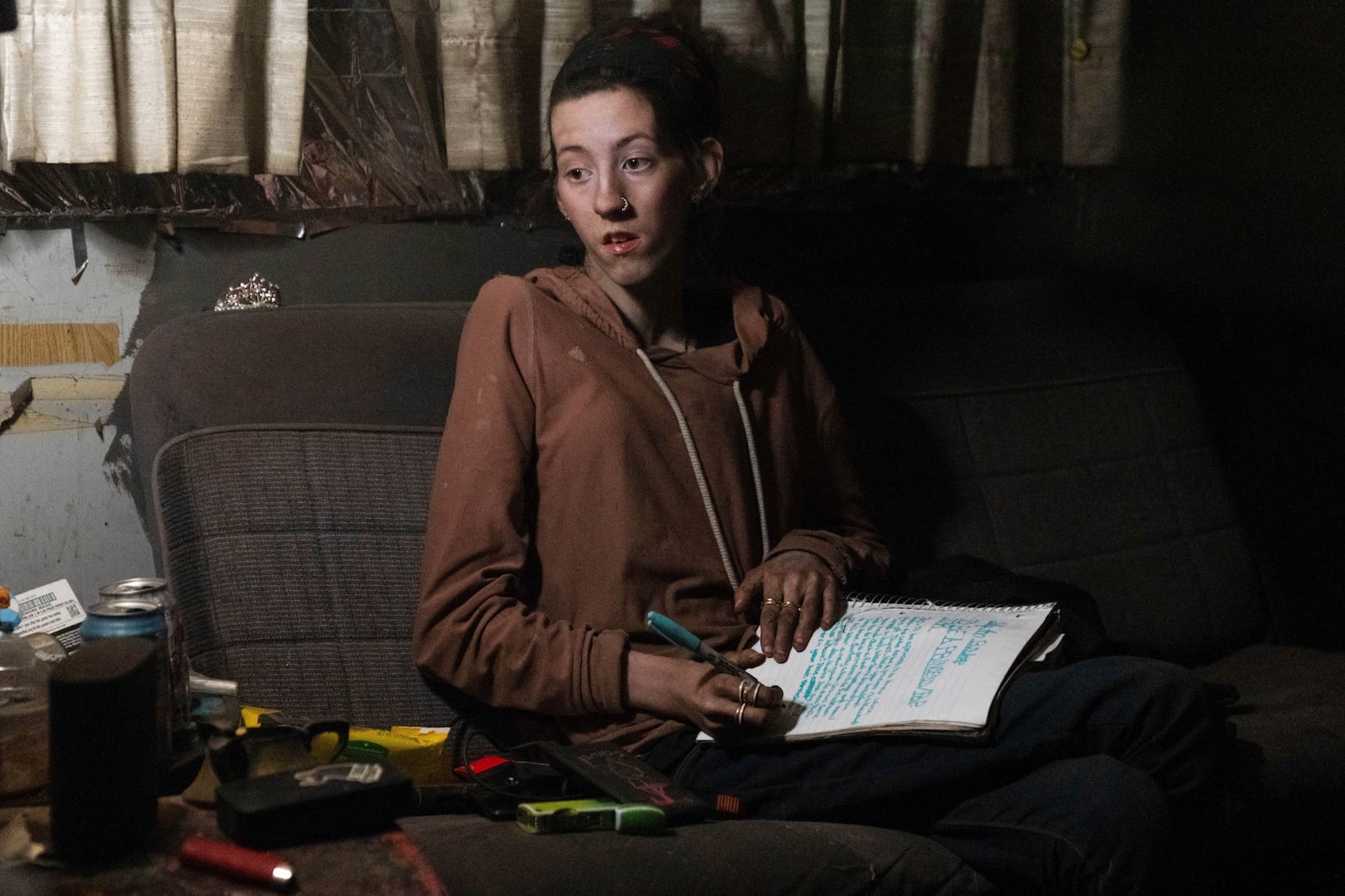 A woman looks up from writing in a notebook with the words "to be a princess" seen near the top of the page and a tiara next to her inside a trailer as her father works to get it to start, at a homeless encampment in Deschutes National Forest, Wednesday, April 30, 2025 near Bend, Ore. (AP Photo/Jenny Kane)
