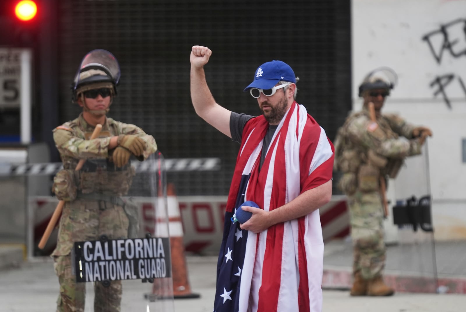 A lone demonstrator draped in a national flag stands outside of the Metropolitan Detention Center, in front of members of the California National Guard, Thursday, June 12, 2025, in Los Angeles. (AP Photo/Damian Dovarganes)