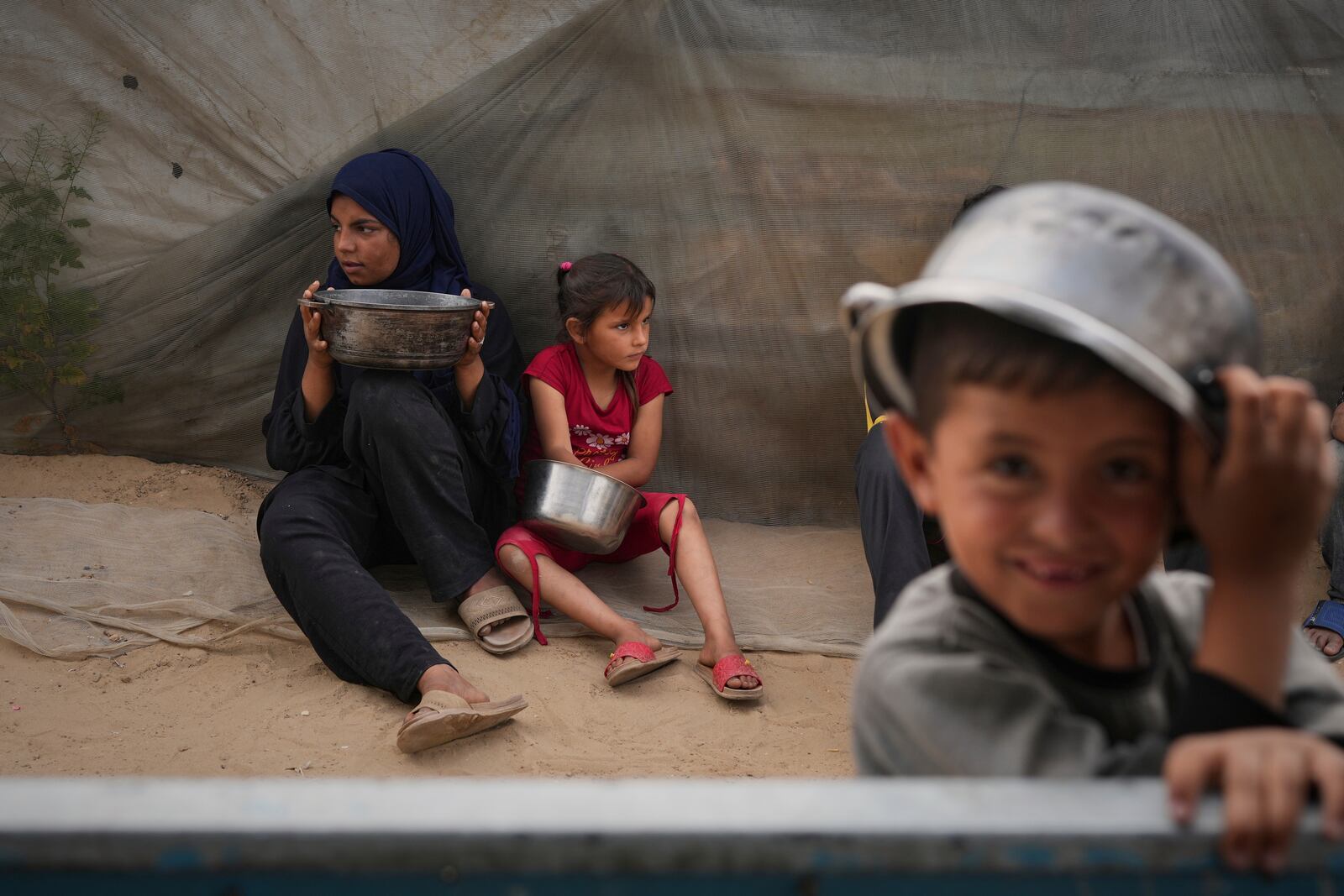 Mira Abu Shaar, 5, right, and her older sister, Raghad, 15, hold pots next to their family tent, as they wait for food to be prepared, in Muwasi, on the outskirts of Khan Younis in the southern Gaza Strip, Thursday, April 24, 2025. (AP Photo/Abdel Kareem Hana)
