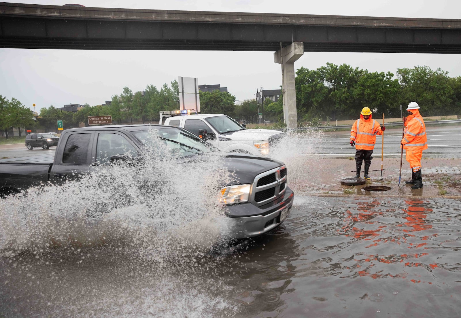 A pick-up truck passes as Tristan Neal, and Kirk Jackson with TxDOT work to clear a clogged sewer drain that caused a N Central Expressway frontage road to flood in Dallas, Wednesday, April 30, 2025. (Juan Figueroa/The Dallas Morning News via AP)