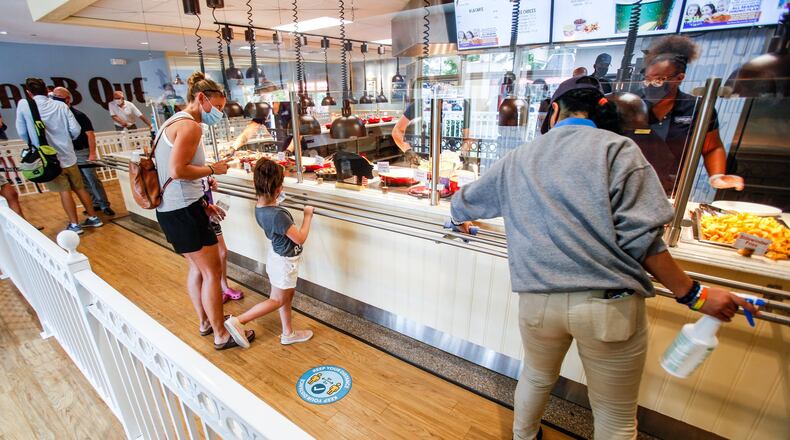 An employee cleans while visitors walk through the food line at Coney Bar B Que on media day at Kings Island Wednesday, July 1, 2020 in Mason. Kings Island opens to pass holders July 2 with numberous protocols in place to decrease the chance of spread of COVID-19. Guest must pre-register for admission to the park and are required to wear masks and stand in accordance with social distancing guidelines. There are also hundreds of hand sanitizers statios around the park. NICK GRAHAM / STAFF