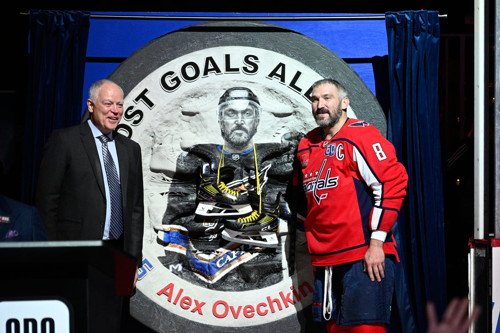 Washington Capitals left wing Alex Ovechkin (8) stands next to one of his gifts and Brian MacLellan, president of Capitals hockey operations, as he takes part in a ceremony to honor him for scoring 895 career NHL goals before an NHL hockey game against the Carolina Hurricanes, Thursday, April 10, 2025, in Washington. (AP Photo/Nick Wass)