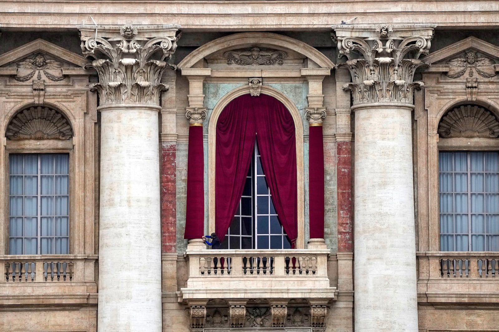 A man gives the last touch to a red drape wrapped around a column of the central lodge of St. Peter's Basilica in Vatican City, Monday, May 5, 2025, before the conclave starting on May 7, where they will elect the 267th pontiff of the Catholic Church. (AP Photo/Gregorio Borgia)