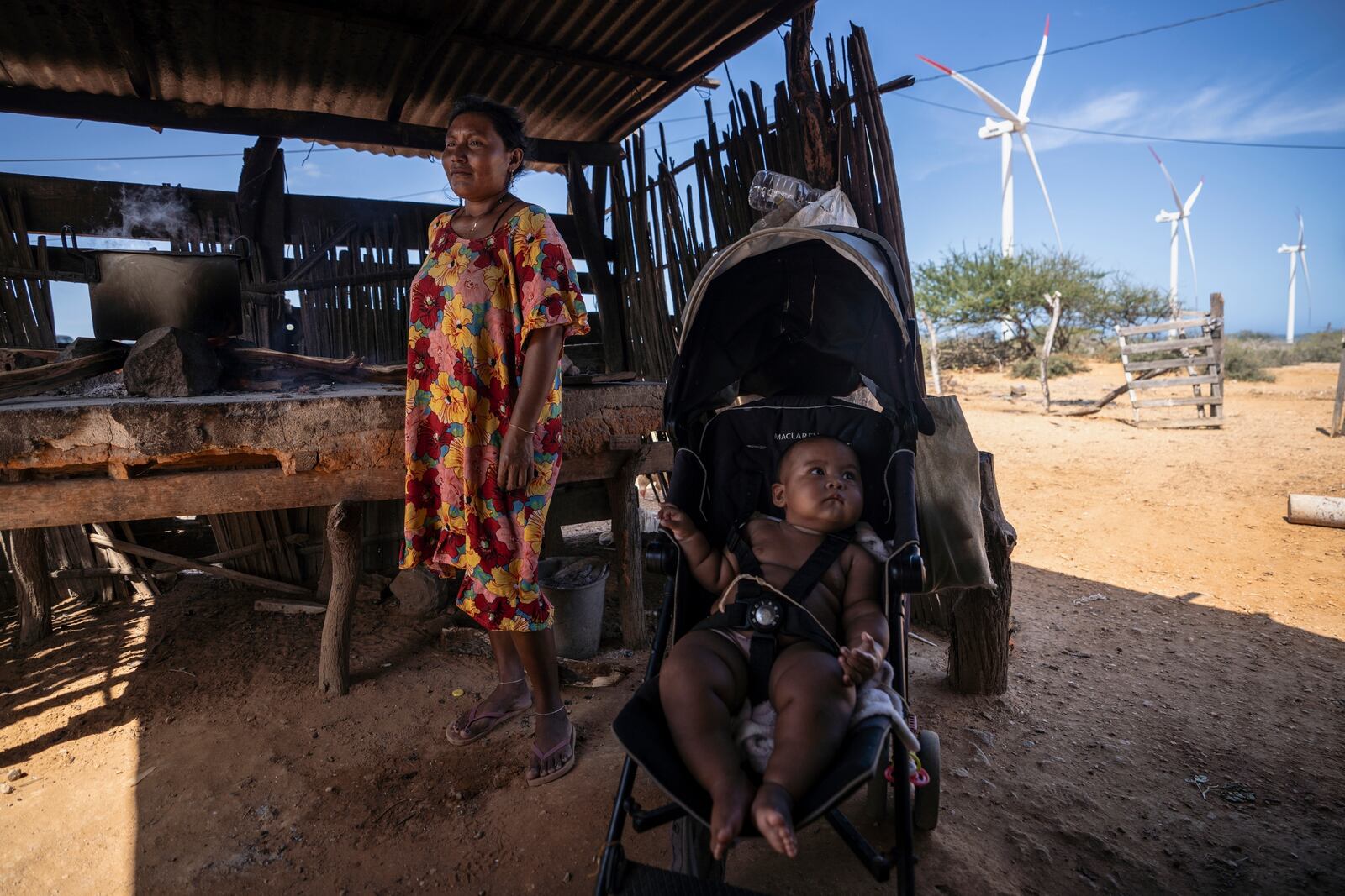 FILE - An Indigenous woman from the Wayuu community cooks near her baby as wind turbines operate in the distance on the outskirts of Cabo de la Vela, Colombia, Feb. 7, 2025. (AP Photo/Ivan Valencia, File)