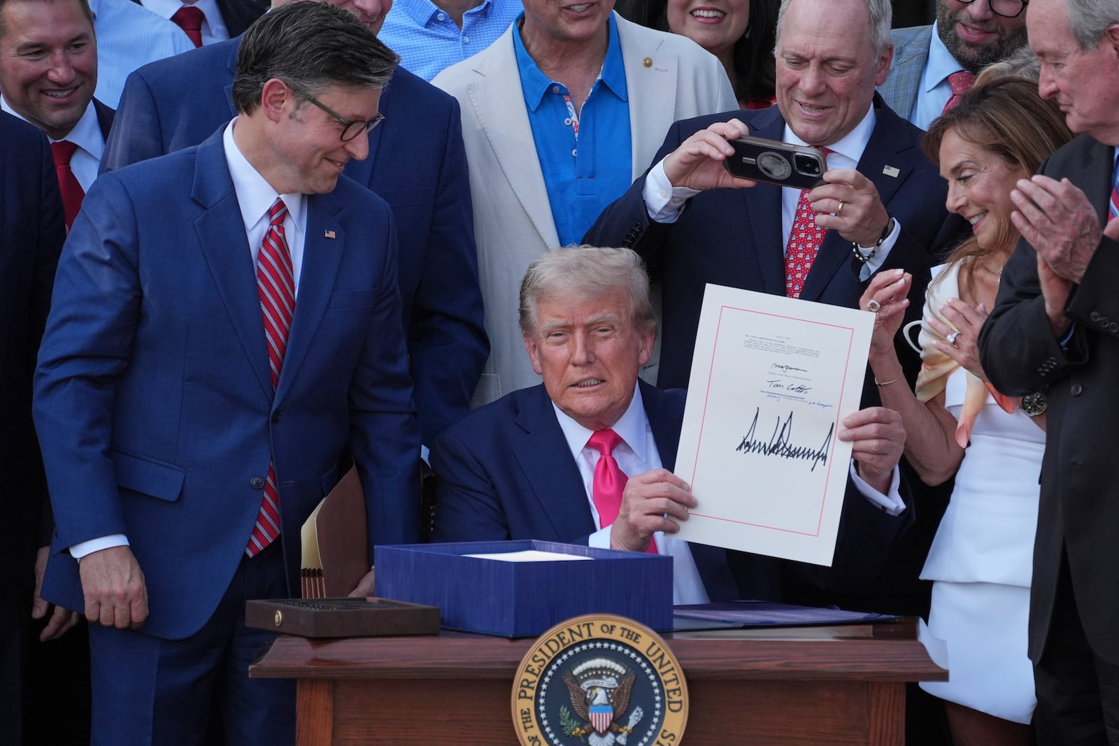 President Donald Trump holds his signed signature bill of tax breaks and spending cuts at the White House, Friday, July 4, 2025, in Washington, as House Speaker Mike Johnson of La., left, watches and Rep. Steve Scalise, R-La., takes a photo. (AP Photo/Evan Vucci)