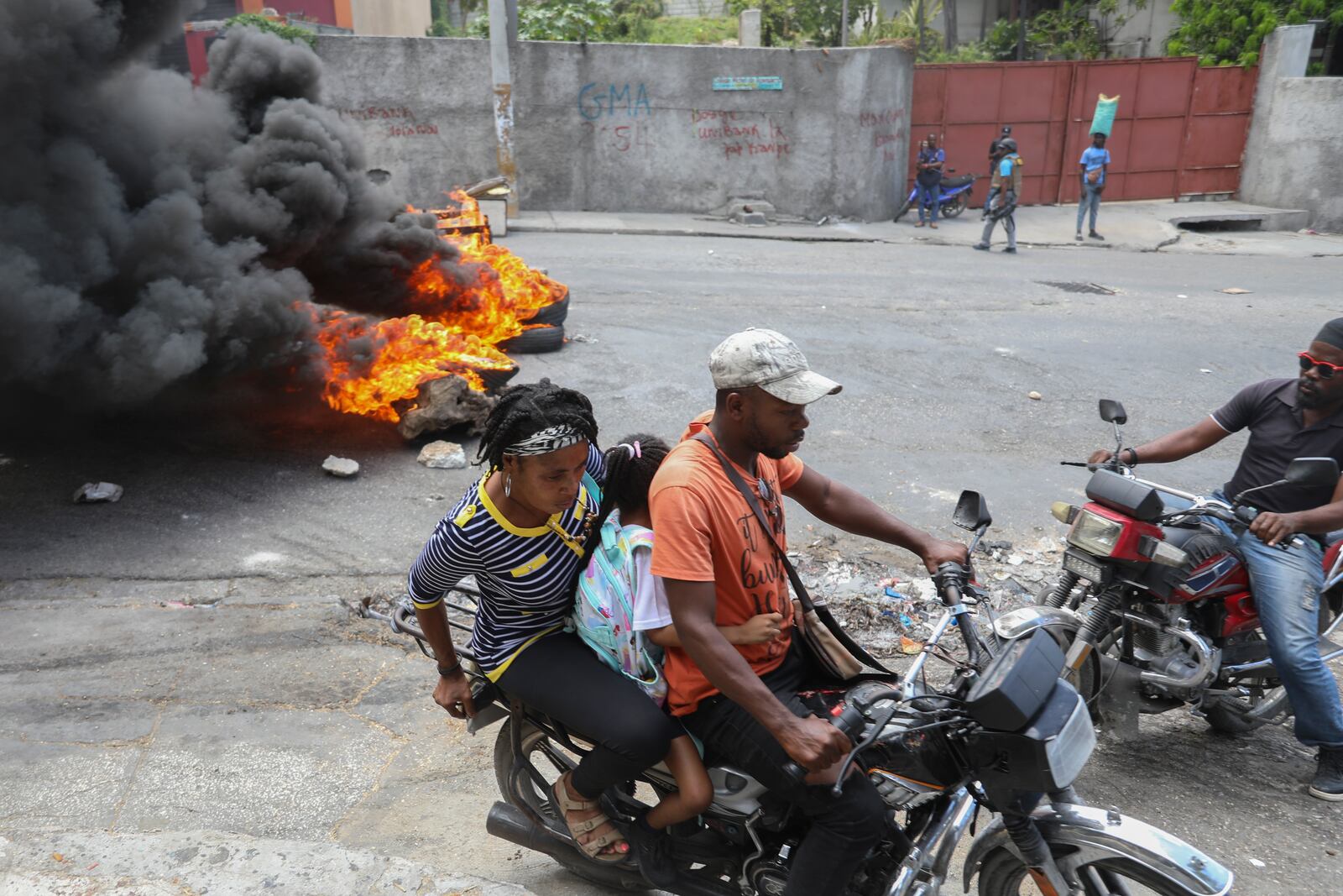 A motorcycle taxi driver transports clients past tires set on fire by protesters during a demonstration demanding the resignation of the transitional presidential council in Port-au-Prince, Haiti, Wednesday, March 26, 2025. (AP Photo/Odelyn Joseph)