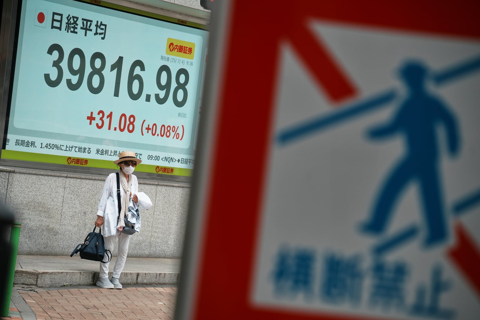 A person seen behind a traffic sign stands in front of an electronic stock board showing Japan's Nikkei index at a securities firm Friday, July 4, 2025, in Tokyo. (AP Photo/Eugene Hoshiko)