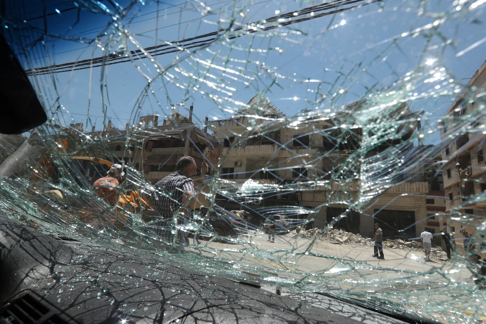 People seen through broken car window, as they gather in front of a destroyed building that was hit in an Israeli airstrike in Nabatieh town, south Lebanon, Friday, June 27, 2025. (AP Photo/Mohammed Zaatari)