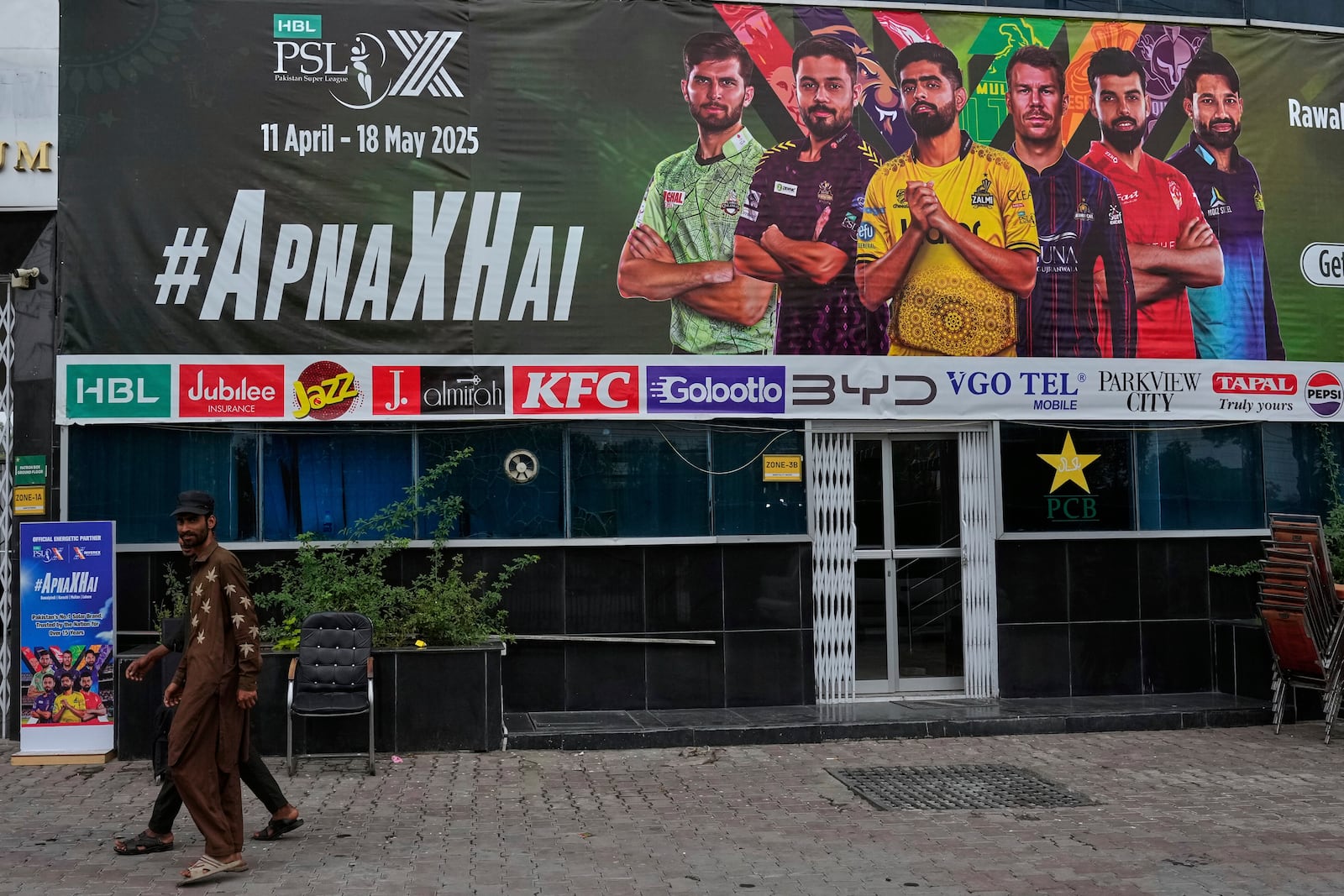 Workers walk past an advertisement board of Pakistan Super League Twenty20 tournament with the pictures of cricketers, installed at the Rawalpindi Cricket Stadium, in Rawalpindi, Pakistan, Friday, May 9, 2025. (AP Photo/Anjum Naveed)