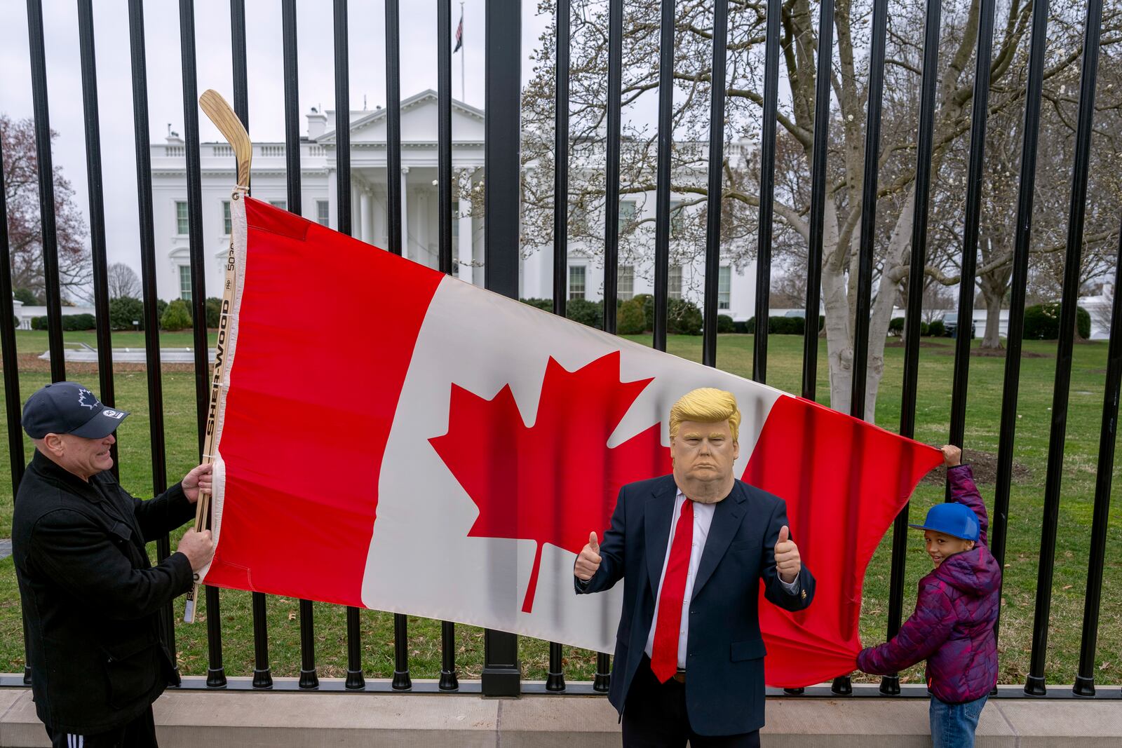 Toronto residents Douglas Bloomfield, from right, and his son Phoenix, who are on vacation in Washington, hold a Canadian flag and an ice hockey stick to show their support for Canada regarding trade tariffs as they pose with with another visitor to the city wearing a mask of President Donald Trump in front of the White House in Washington, Thursday, March 13, 2025. (AP Photo/Ben Curtis)
