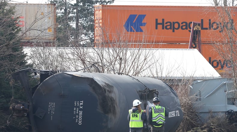 A HAZMAT team member and an Ohio EPA representatives stand by an overturned tank car as they watch another train travel down the tracks in the background Monday, March 6, 2023, in Springfield. A Norfolk Southern train derailed in Clark County near the fairgrounds on Saturday. BILL LACKEY/STAFF
