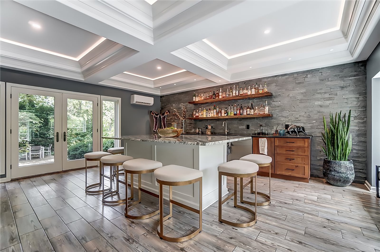 The first floor entertainment room has an island with bar seating, a coffered ceiling and recessed lighting, an island with a quartz top and built in cabinets with a sink and beverage coolers. There is a stacked stone accent wall and French doors that open to the back paver patio. CONTRIBUTED