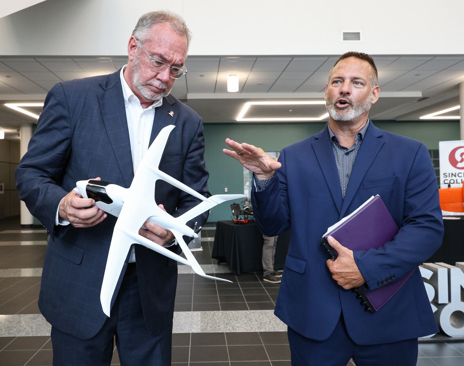 Sinclair Community College Senior Vice President Jeffery Miller talks on Wednesday, Aug. 13 at Ned J. Sifferlen Health Sciences Center as President Steven Johnson holds the model of an aircraft the college is purchasing. The aircraft is expected to be delivered in early 2026. BRYANT BILLING / STAFF