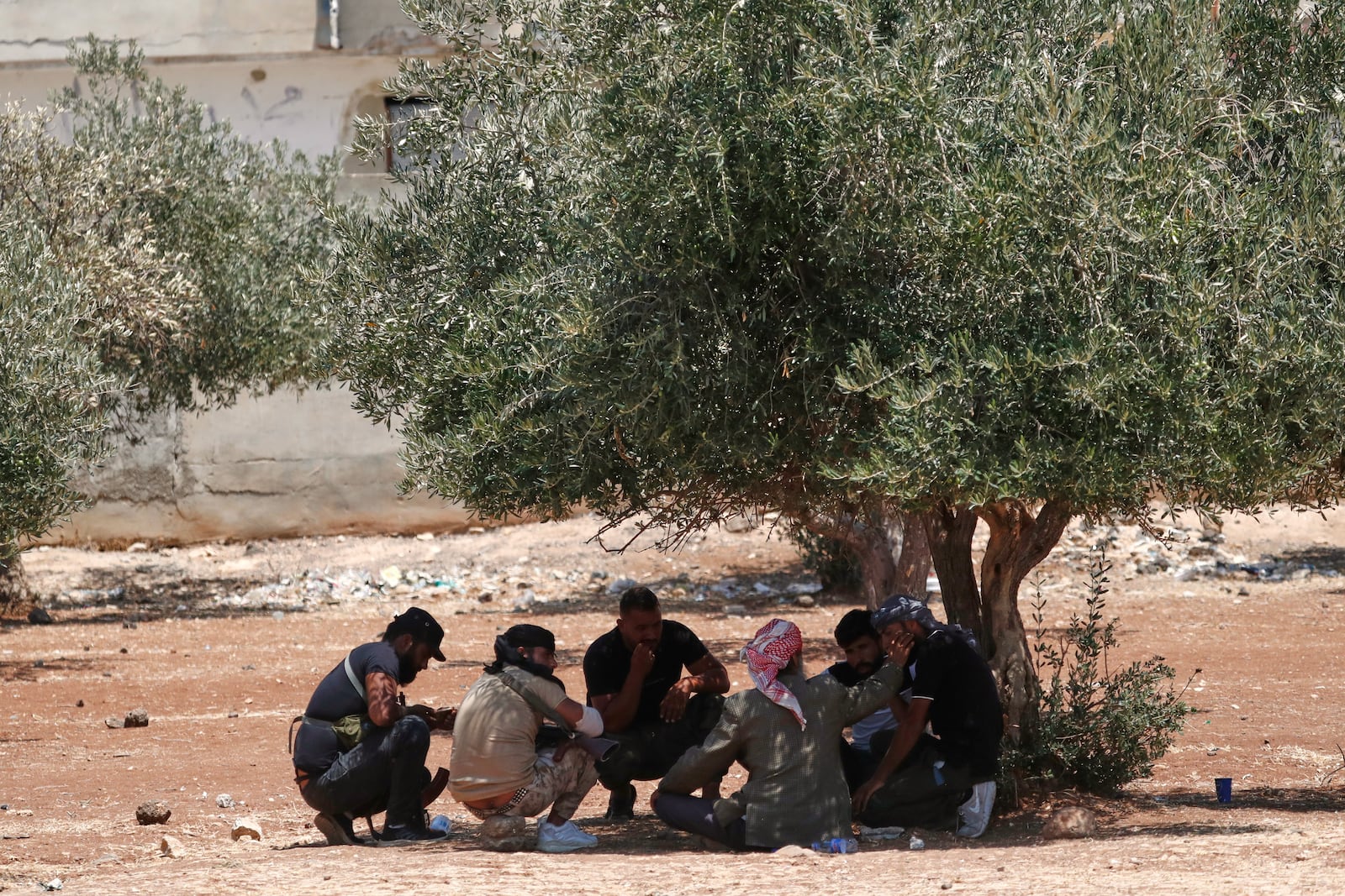 Bedouin fighters sit under the shade of an olive tree outside a temporary shelter housing Bedouin families who fled their homes in Shahba, a town in Syria's southern Sweida province, due to clashes between Bedouin clans and Druze militias, in the town of Nahtah, in the eastern part of Daraa province, Syria, Monday, July 21, 2025. (AP Photo/Omar Sanadiki)