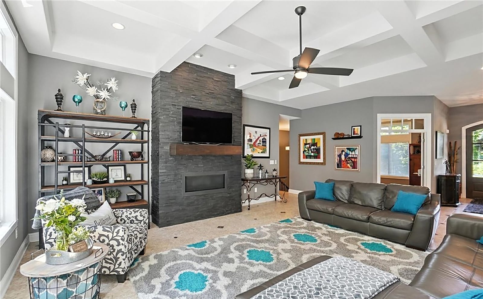 The great room has a stone fireplace, coffered ceiling, tile floor, recessed lighting and a ceiling fan.