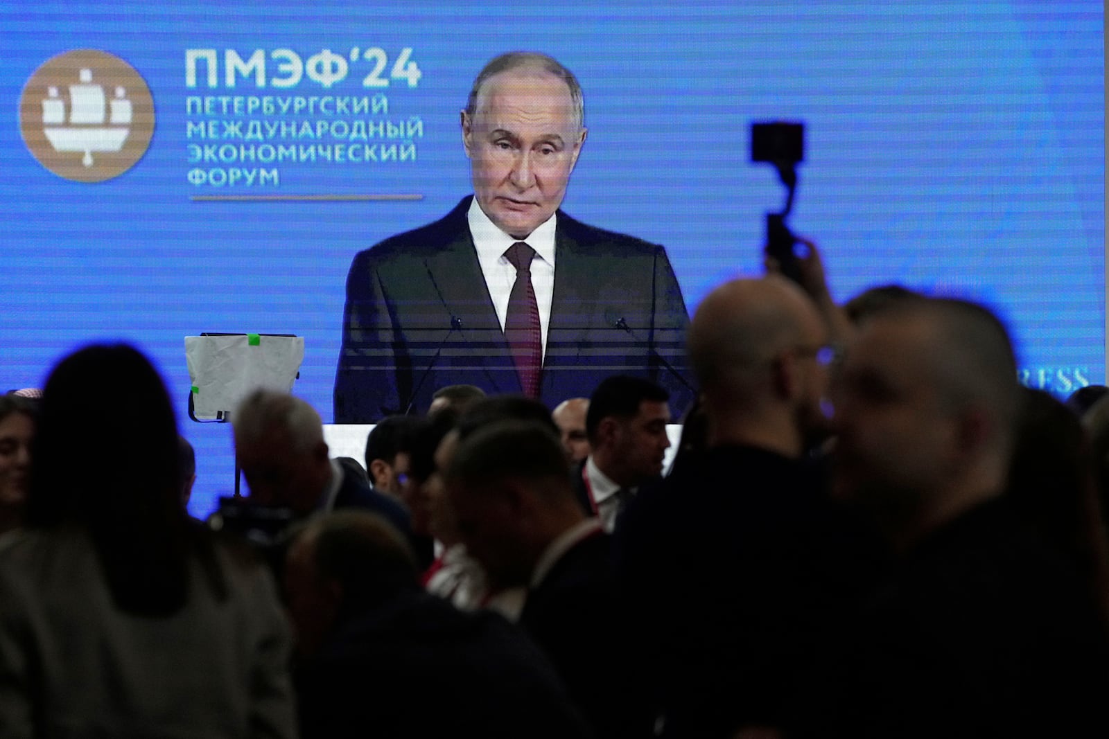 Guests and participants walk in front of a TV screen showing Russian President Vladimir Putin on the sidelines of the St. Petersburg International Economic Forum in St. Petersburg, Russia, Wednesday, June 18, 2025. (AP Photo/Dmitri Lovetsky)