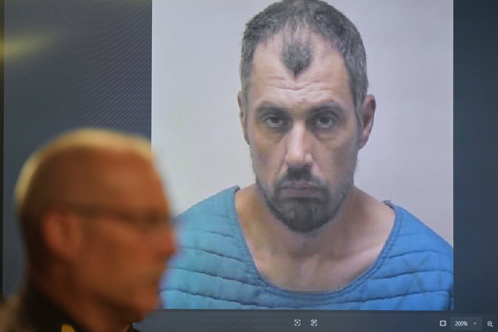 The booking photo of Bradford Gille, who is accused of stabbing 11 people at a Walmart, is seen behind Grand Traverse County Sheriff Mike Shea during a news conference Sunday, July 27, 2025, in Traverse City, Mich. (AP Photo/Ryan Sun)