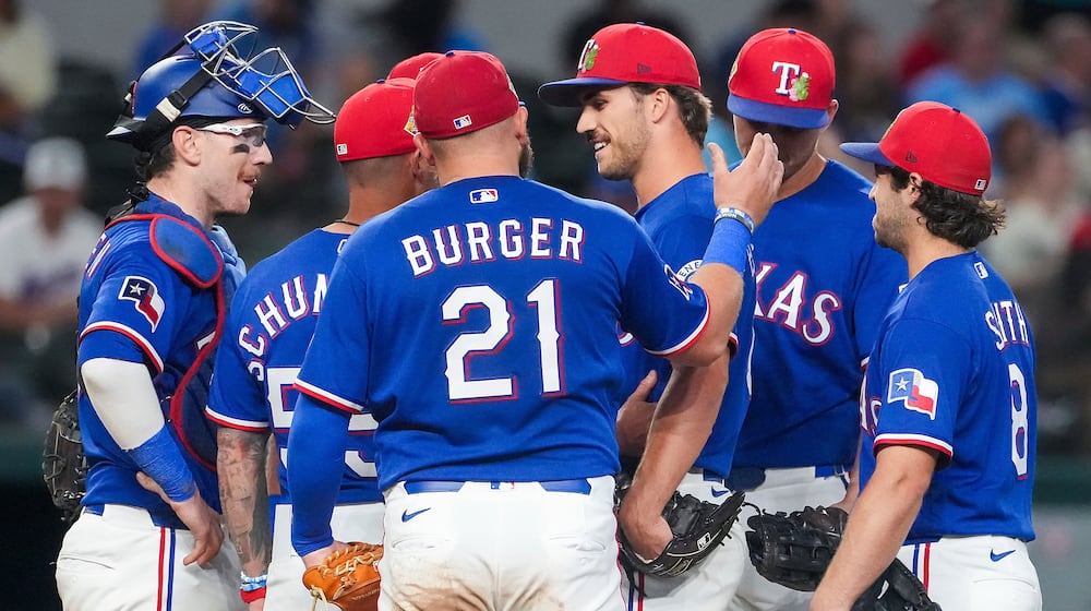 Texas Rangers pitcher Carter Baumler, third from right,gets a pat on the back from first baseman Jake Burger (21) as he gets a visit at the mound from manager Skip Schumaker, second from left, to inform him he has made the opening day roster during the fifth inning of an exhibition baseball game against the Kansas City Royals, Monday, March 23, 2026, in Arlington, Texas. (Smiley N. Pool/The Dallas Morning News via AP)